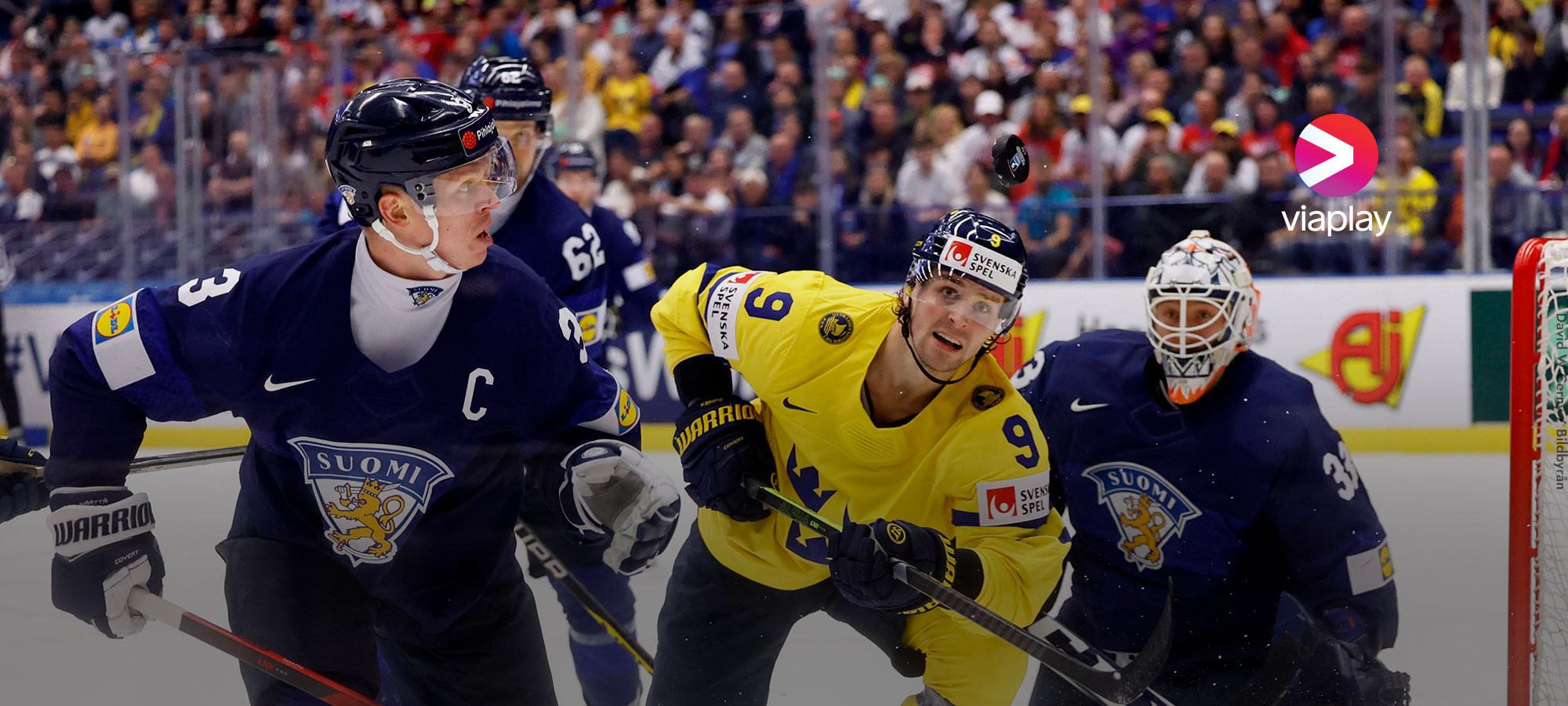 Kuvassa on Suomi - Ruotsi jääkiekko-ottelu. Kuvassa näkyy kolme Suomen pelaajaa ja yksi Ruotsin pelaaja. Yksi Suomen pelaajista on maalivahti, joka on polvillaan jäällä. Oikeassa yläkulmassa Viaplay-logo.
