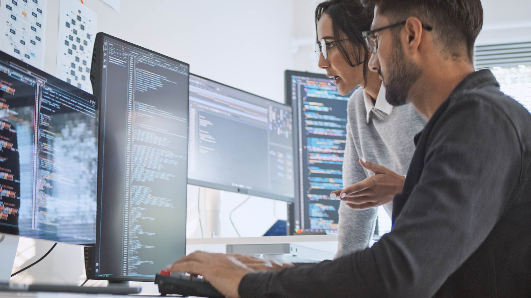 Kaksi IT-asiantuntijaa työskentelee useiden näyttöjen äärellä analysoiden ohjelmakoodia ja tietojärjestelmiä.