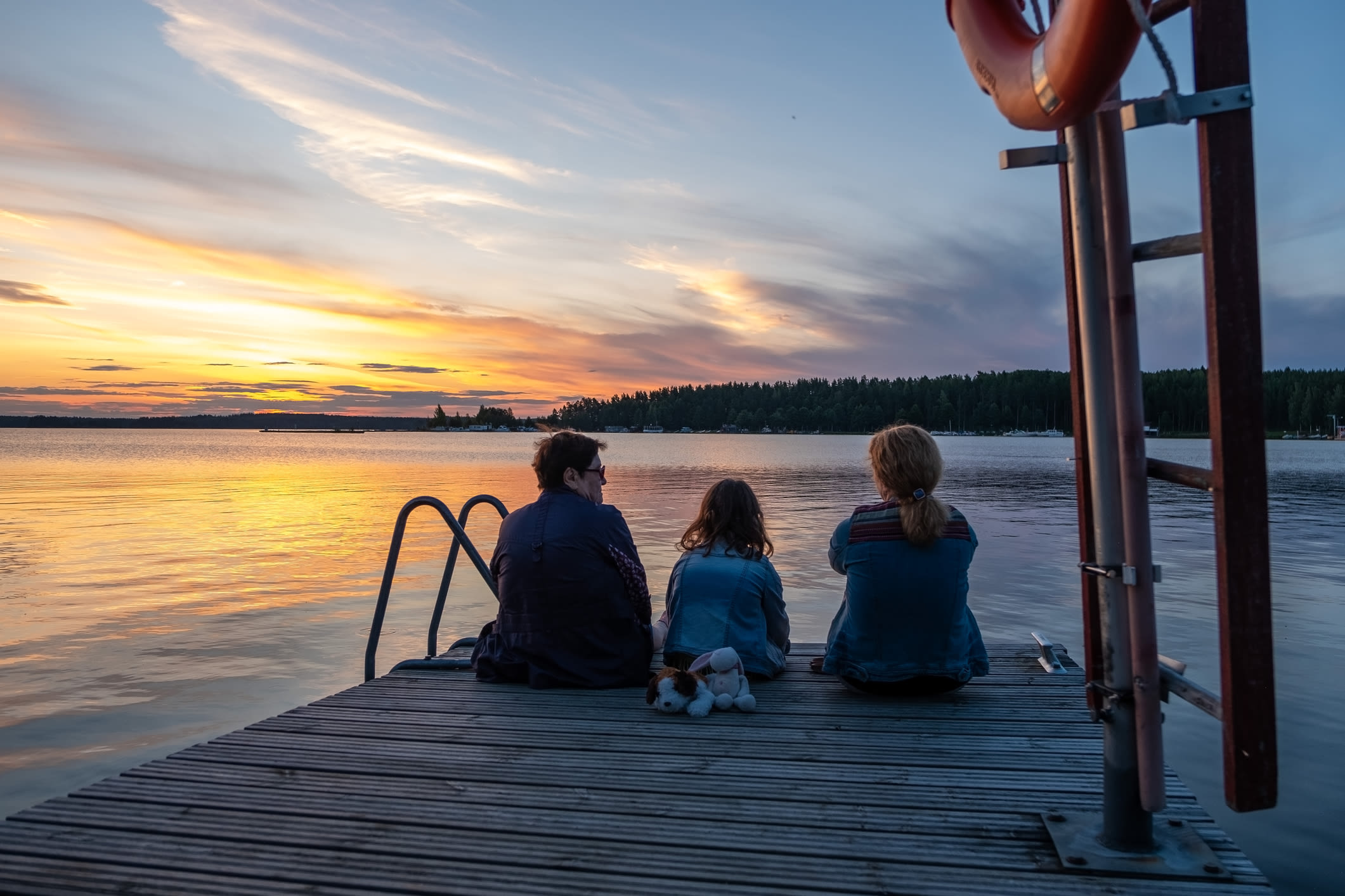 Kolme henkilöä istuu mökkilaiturilla auringonlaskua katsellen