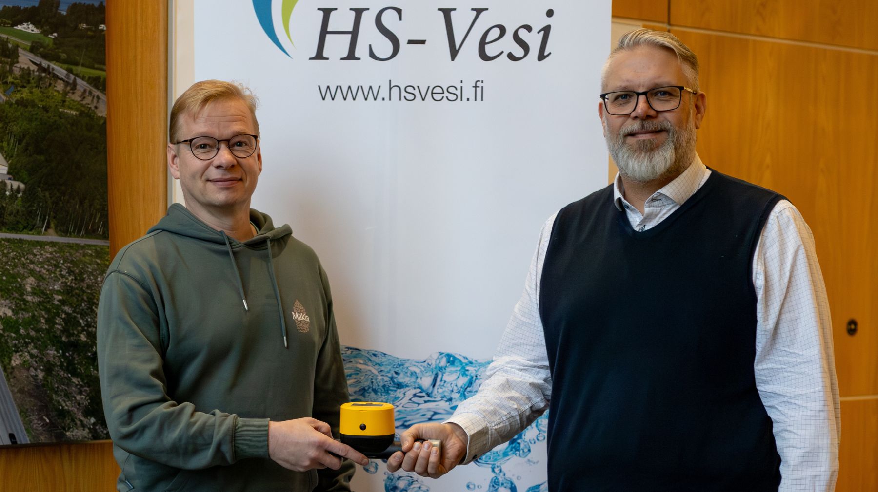 Elisan ja HS-Veden edustajat poseeraavat kameralle ja pitelevät yhdessä keltaista vesimittaria. Taustalla näkyy HS-Vesi -logolla varustettu roll-up ja seinäkuvia vesihuoltoon liittyvistä maisemista.