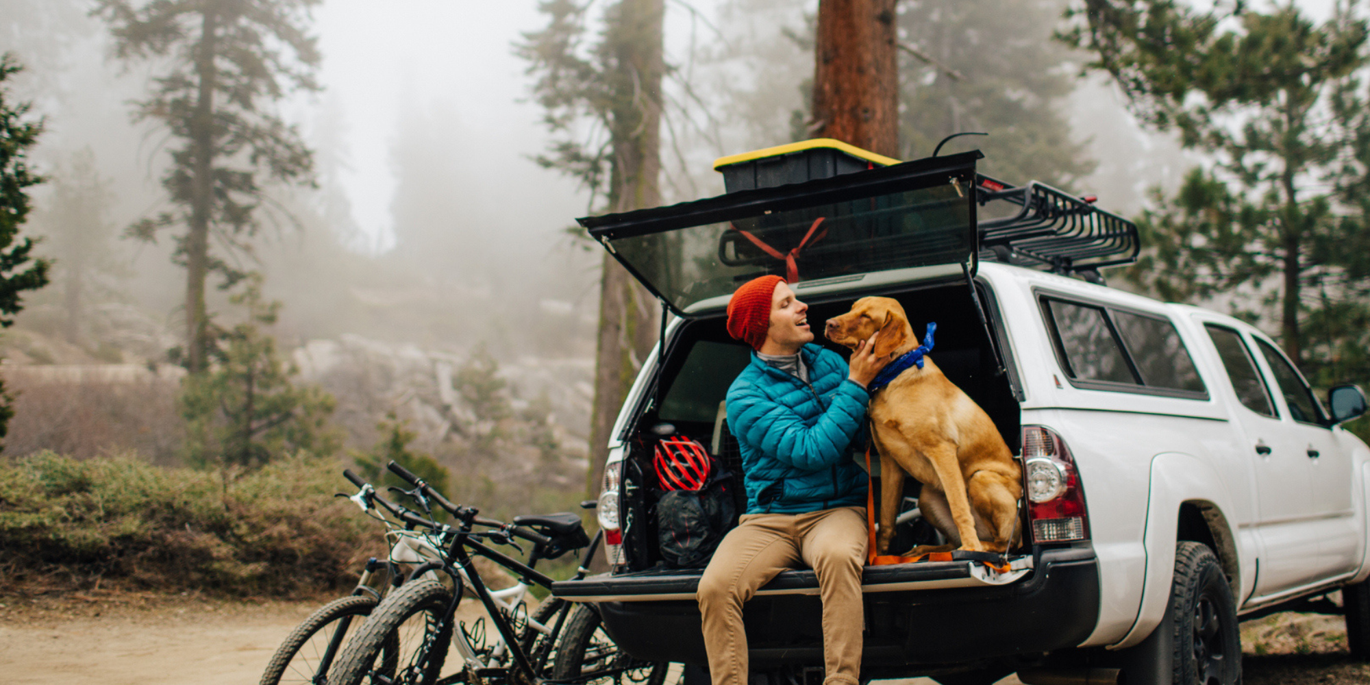 mies ja koira istumassa maastoauton takaluukun päällä, vieressä kaksi maastopyörää