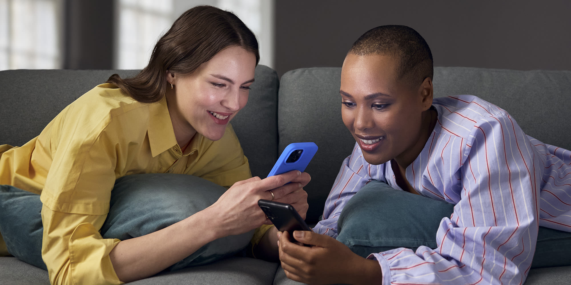 nuori mies ja nainen makaavat vatsallaan sohvalla kasvot toisiaan kohti, molemmilla on kuulokkeet korvilla, puhelimet kädessä ja he katsovat toisiaan hymyillen.