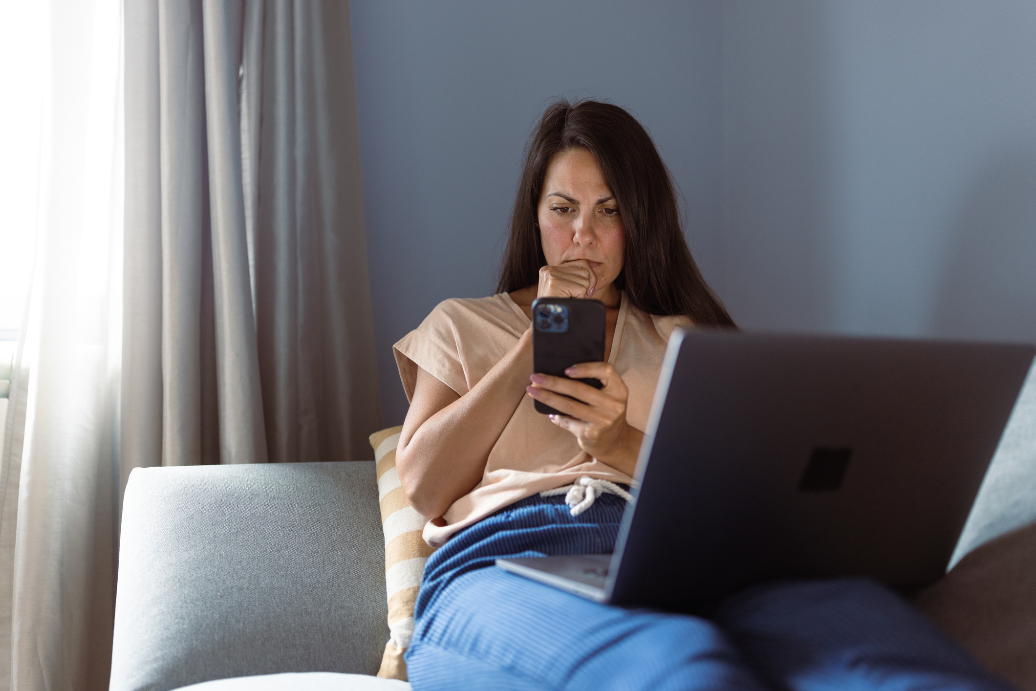 Nainen istuu sohvalla läppäri sylissään ja katsoo puhelintaan huolestuneena