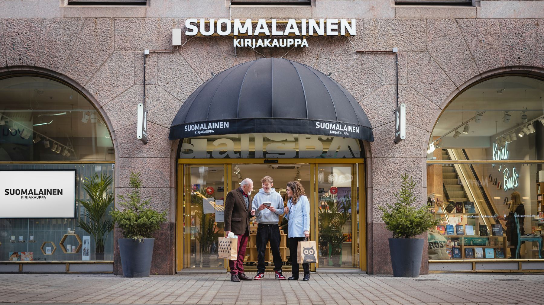 Vanhempi mies, nainen ja poika seisovat ulkona Suomalaisen Kirjakaupan sisäänkäynnin edessä.
