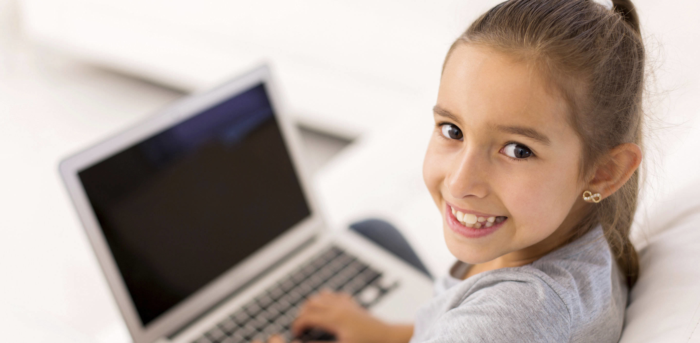 tummatukkainen ja ruskeasilmäinen kouluikäinen tyttö istuu pöydän ääressä ja katsoo kameraan. Edessään hänellä on läppäri.