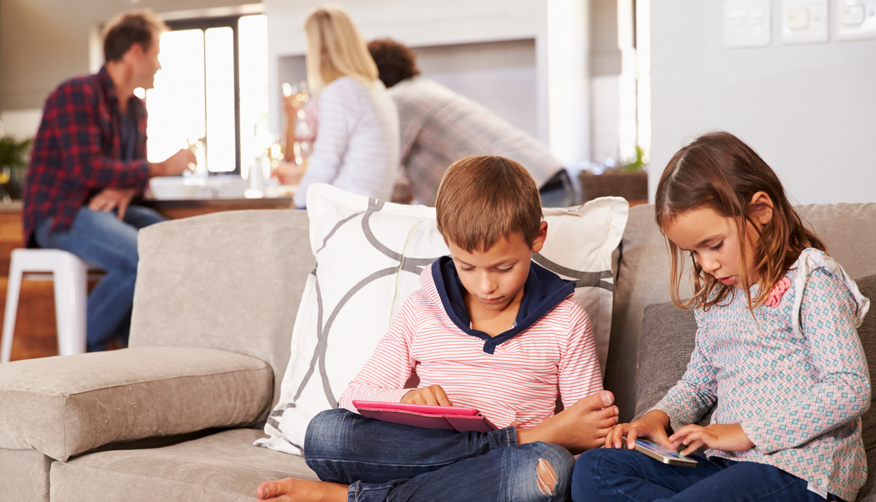 tyttö ja poika istuvat sohvalla ja pelaavat tableteilla. Taustalla aikuiset juttelevat keskenään keittiön pöydän ääressä.