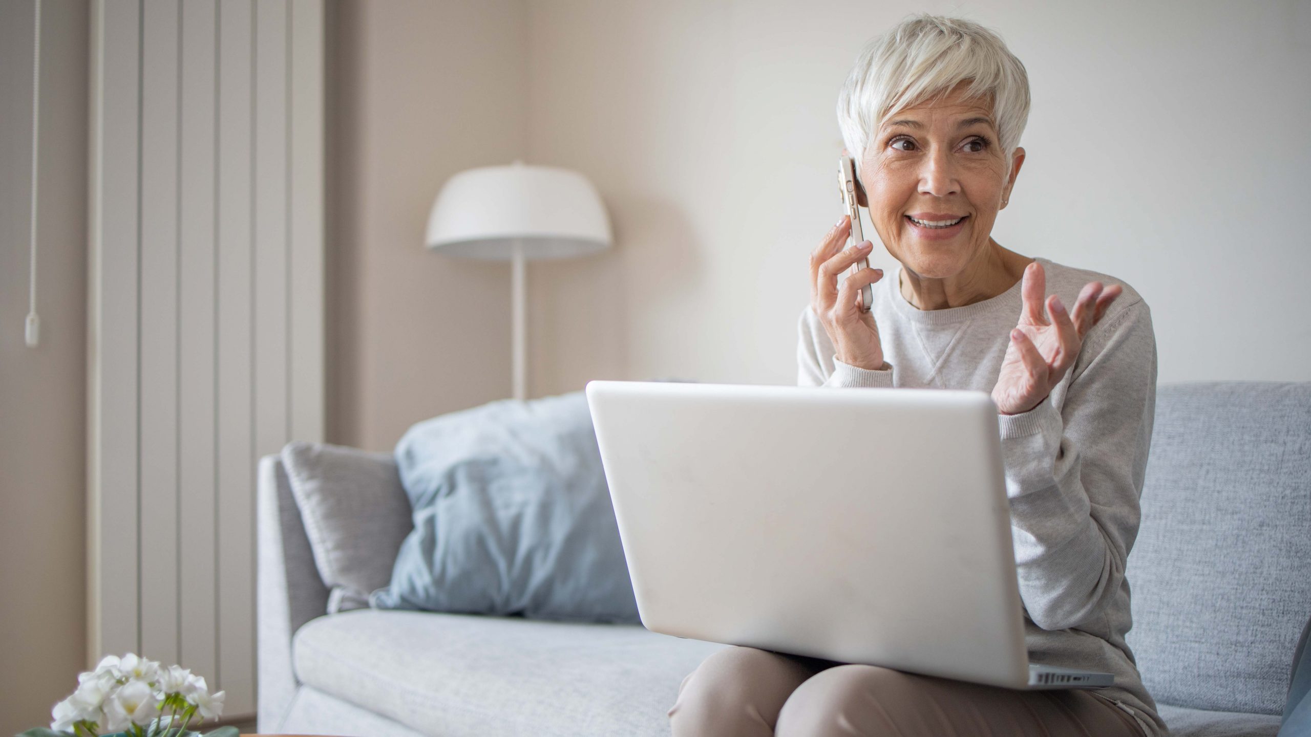 Harmaahiuksinen nainen istuu sohvalla läppäri sylissään ja puhuu samalla puhelimeen