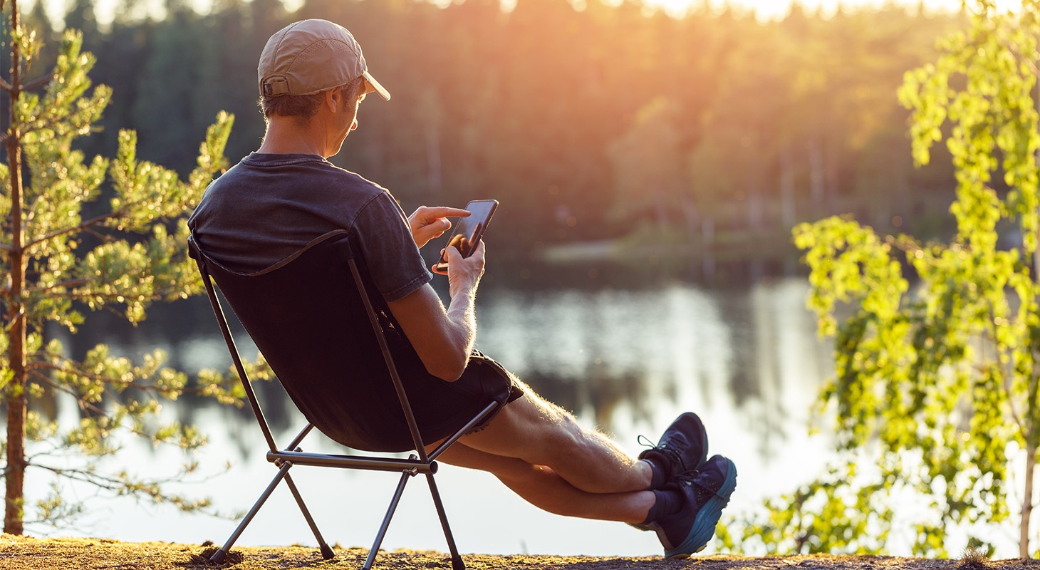 Henkilö istuu kesällä vede äärellä retkituolissa ja selailee älypuhelinta.