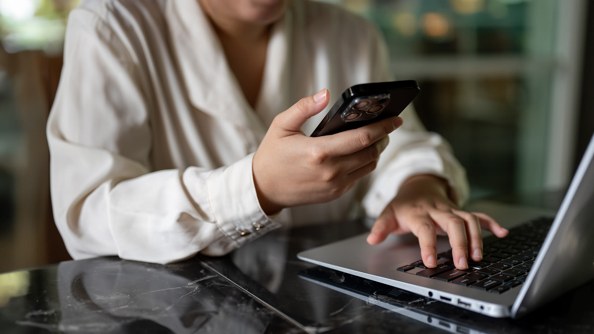 kuva naisesta työpöydän ja läppärin ääressä, kädessä naisella iPhone.