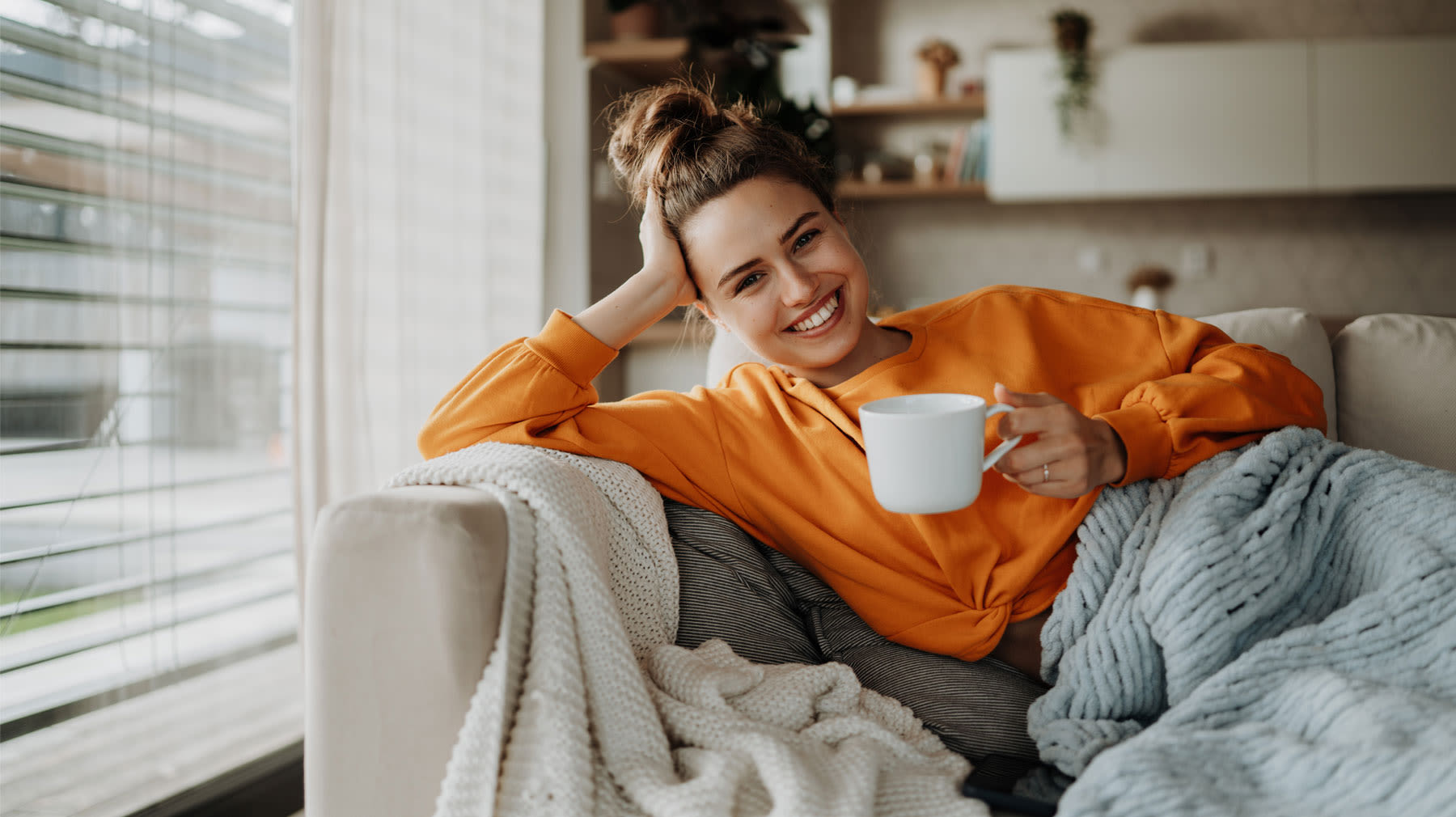 Hymyilevä nainen istuu sohvalla kahvikuppi kädessään. Mielen rauhoittaminen.