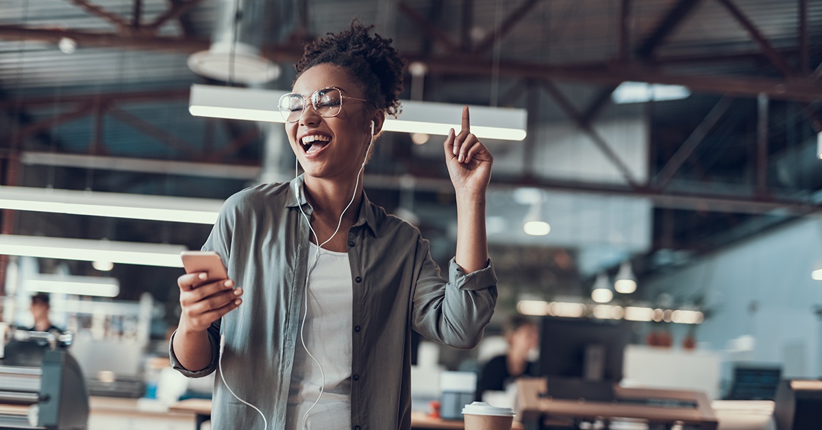 Mitä kaikkea moderni yritysverkko tarjoaa? Iloinen ja naurava afroamerikkalainen nainen nappikuulokkeet päässä katselee matkapuhelimensa näyttöä