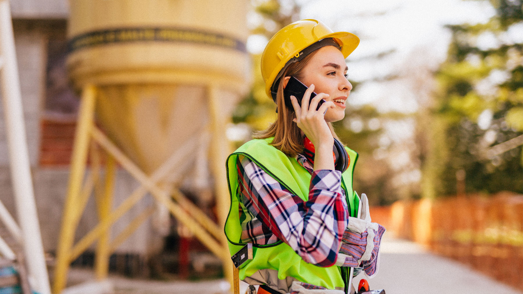 Nainen puhuu puhelimeen rakennustyömaalla. Hänellä on suojaliivi ja kypärä.