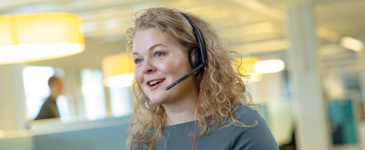 Kiharahiuksinen asiakaspalvelija hymyilee kuulokkeet korvilla, asiakaspalvelun tehostaminen tekoälyllä tukee työtä.