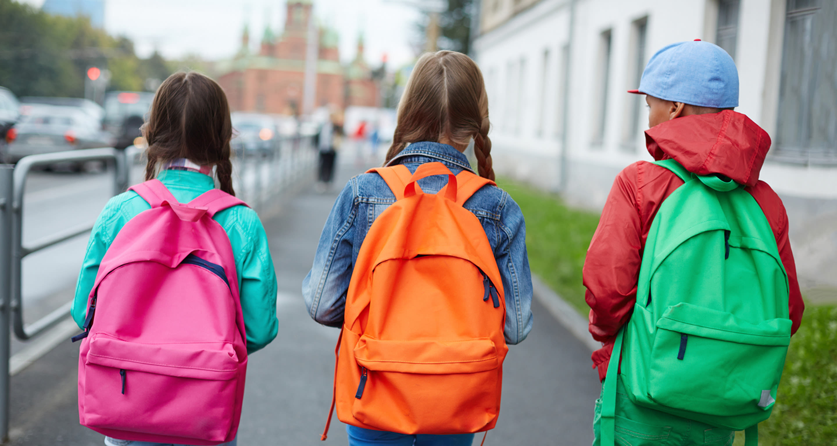 kolme lasta vierekkäin kadulla selät kuvaajaan päin. Kaikilla on selässään eri väriset reput. Vasemman puoleisella tytöllä pinkki, keskimmäisellä oranssi ja oikeassa reunassa olevalla pojalla vihreä.