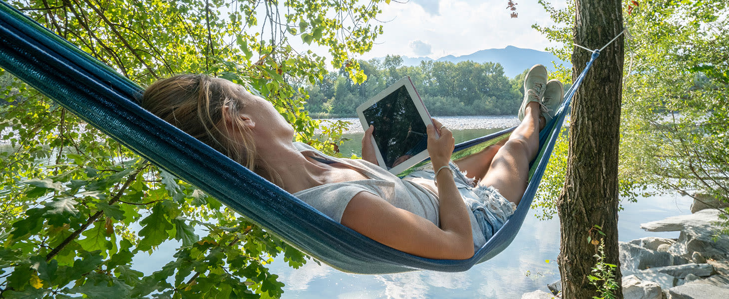 Nainen lukee kirjaa riippumatossa järven rannalla.