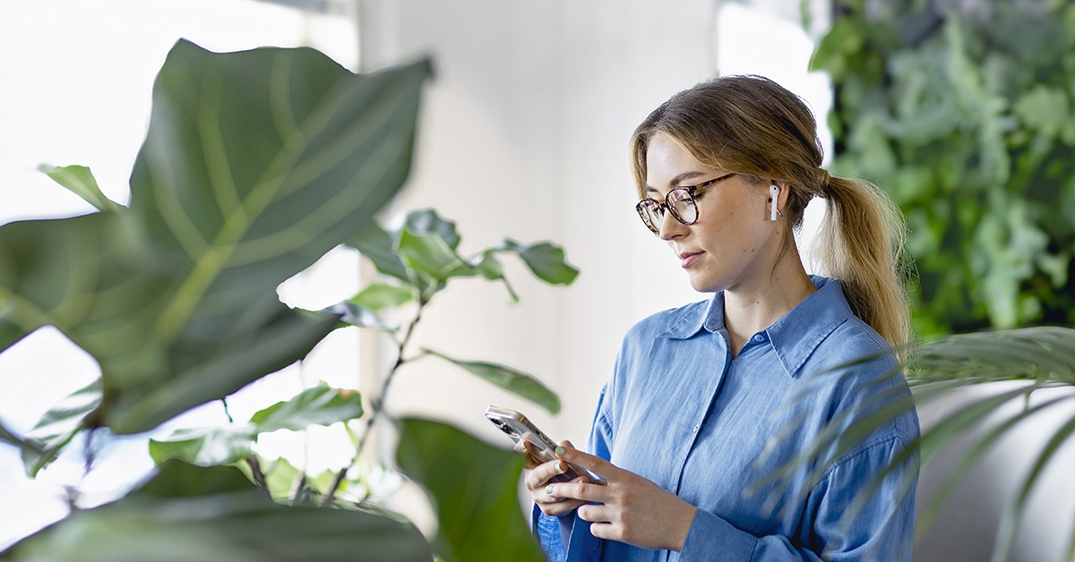 Nainen katselee älypuhelintaan viherkasvien keskellä. Vastaaja lomalla.