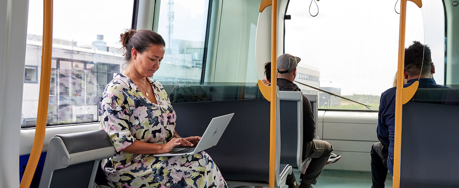 Nainen kesämekossa istuu metrossa ja kirjoittaa tietokoneella.