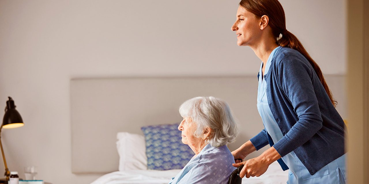 Nainen työntää pyörätuolissa istuvaa vanhusta huoneessa, jossa on sänky ja yöpöydällä valaisin.