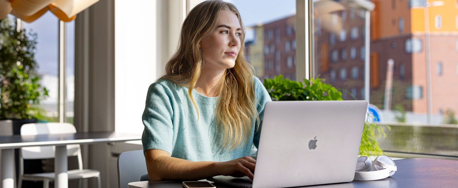 Nainen istuu julkisessa tilassa pöydän ääressä kannettavan tietokoneen edessä ja katselee ikkunasta ulos.