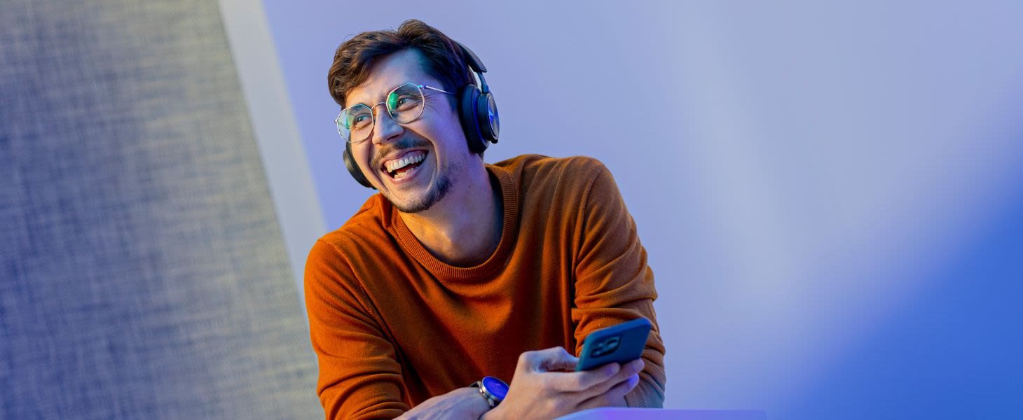 Mies kuulokkeet päässä ja puhelin kädessä katsoo sivulle ja hymyilee sinistä taustaa vasten, suunnitelmissa uusi tekoälyratkaisu.