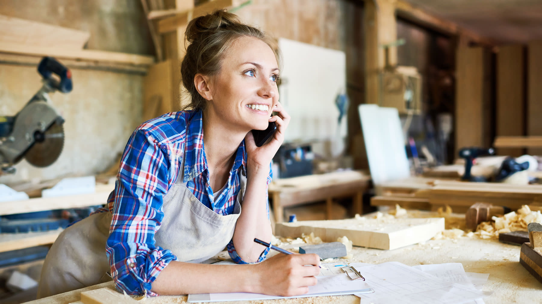 Hymyilevä nainen puhuu puhelimeen ja nojaa pöytään puusepän verstaassa.