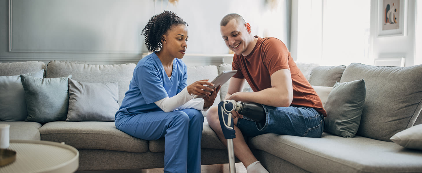 Kuvassa hoitaja ja potilas, jolla on jalkaproteesi, istuvat sohvalla ja katsovat tabletin ruutua.