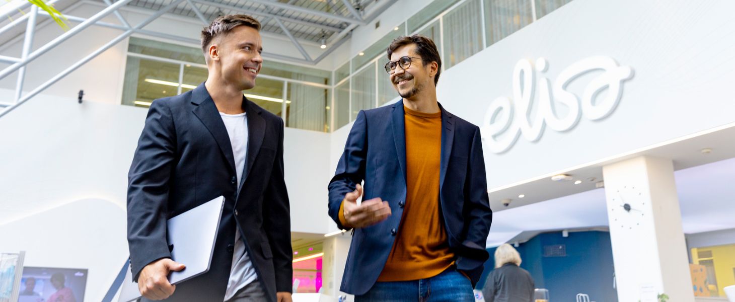 Kaksi miestä kävelemässä toimistolla, Elisan logo taustalla. Tekoälytrendit keskustelun aiheena.